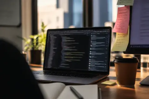 Foto editorial en primer plano del escritorio de un desarrollador, con la pantalla del portátil mostrando un editor de código desenfocado junto a una ventana de chat, notas adhesivas de colores en el borde del monitor con nombres de comandos escritos a mano, luz de fin de tarde