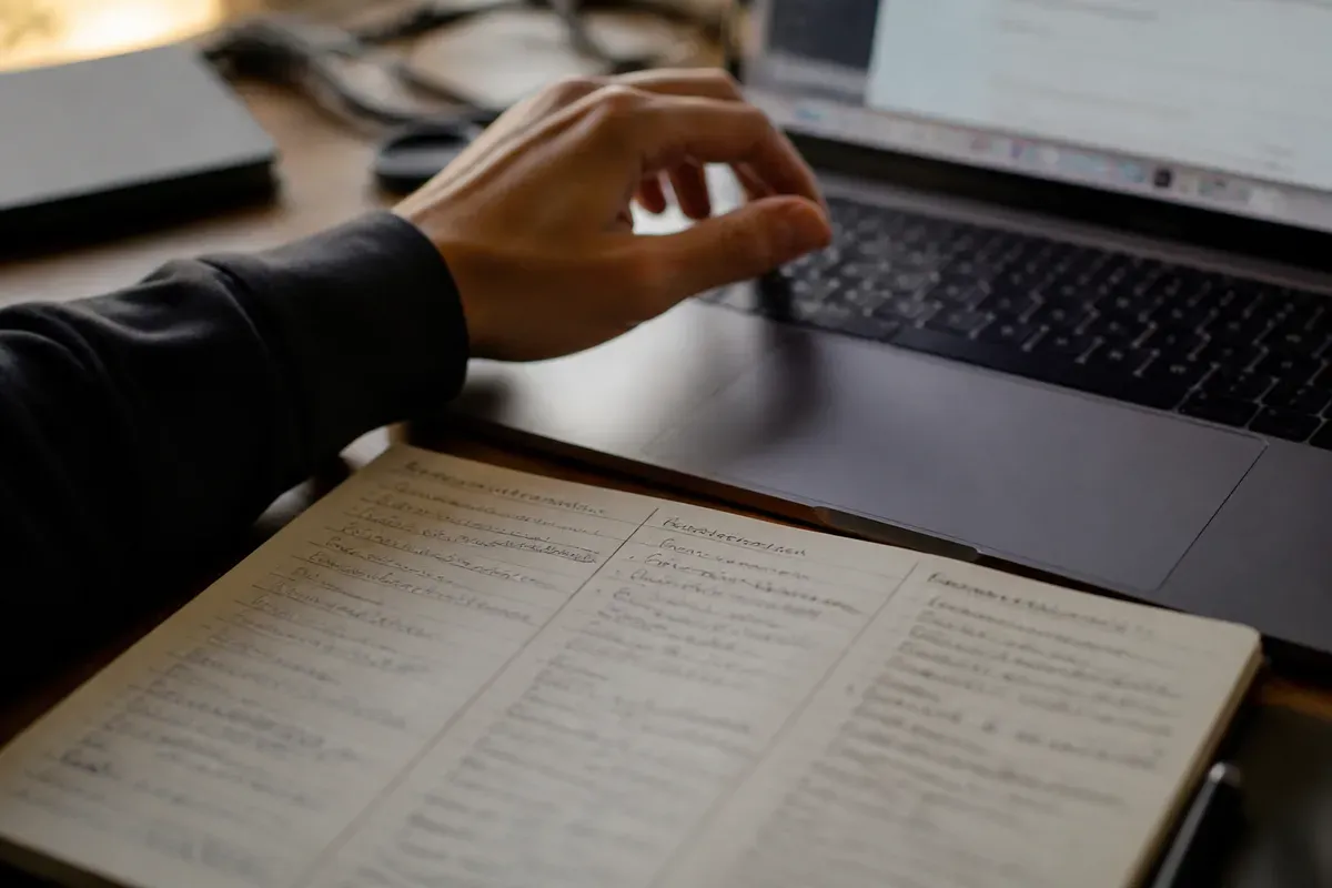 Primer plano de una mano sobre el teclado de un portátil junto a un bloc de notas con tres columnas de anotaciones, con luz suave de fin de tarde